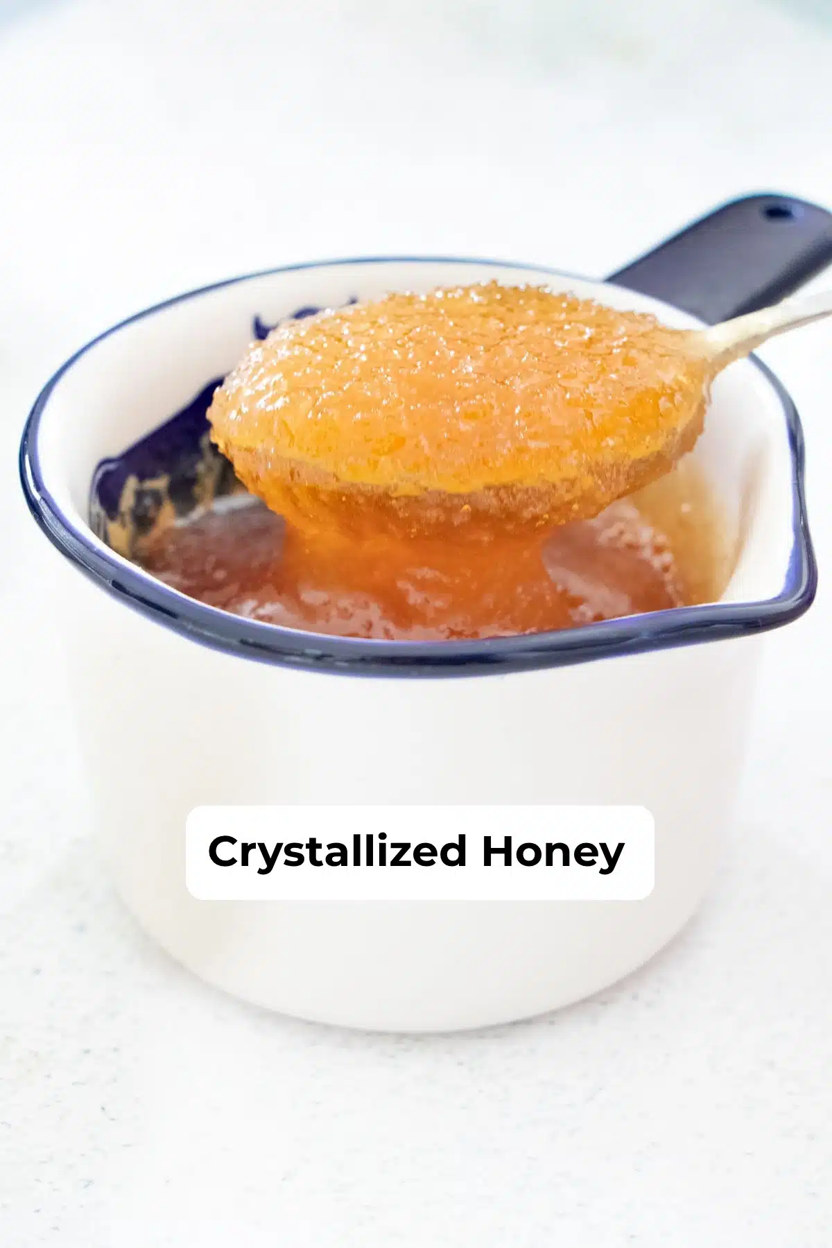 Close up of crystallized, gritty honey on a spoon over a ceramic measuring cup.