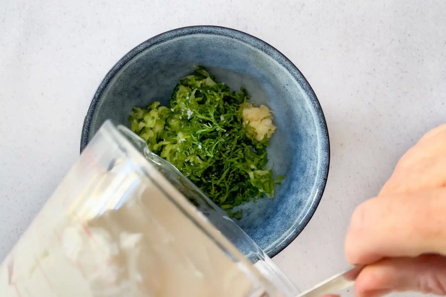A white hand is pouring yoghurt over dill, cucumber, and garlic in a bowl to stir and make into tzatziki.