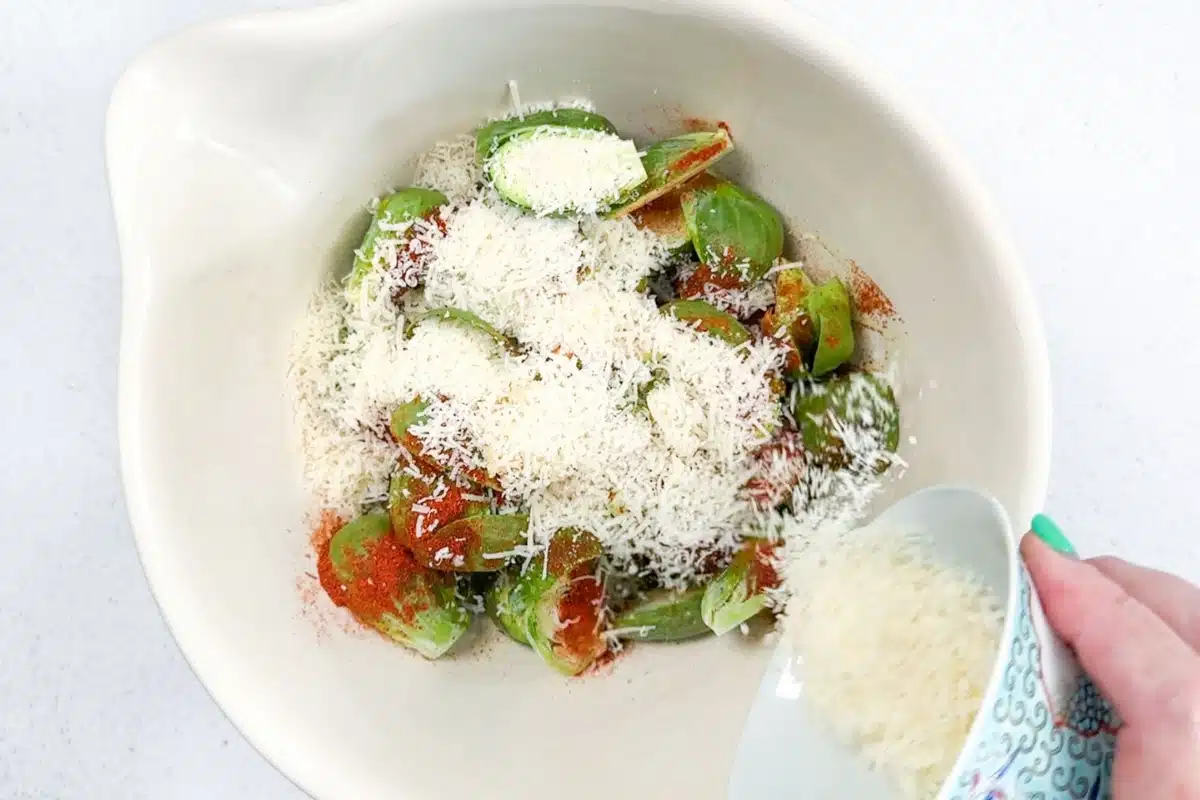 Parmesan cheese being poured into a bowl with Brussels sprouts, oil, and seasonings.