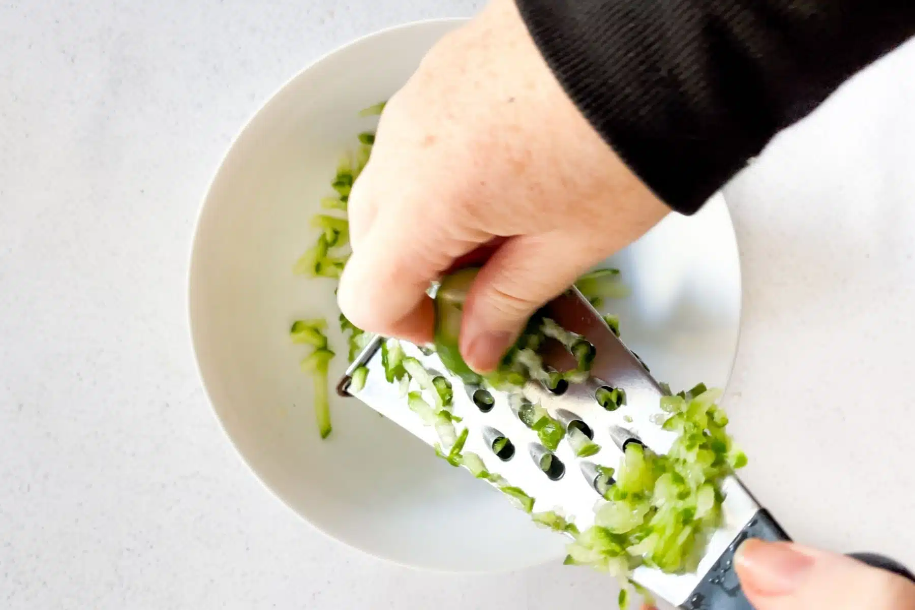 A white hand is using a box grater to grate cucumber into a bowl for tzatziki dip.
