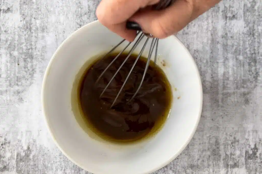 A white hand is mixing a salad dressing for the peach arugula salad with a whisk in a white bowl.