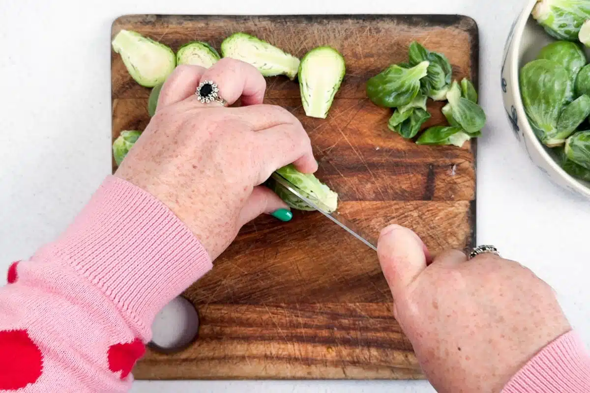 Two white hands are chopping Brussels sprouts in half on a wooden chopping board.