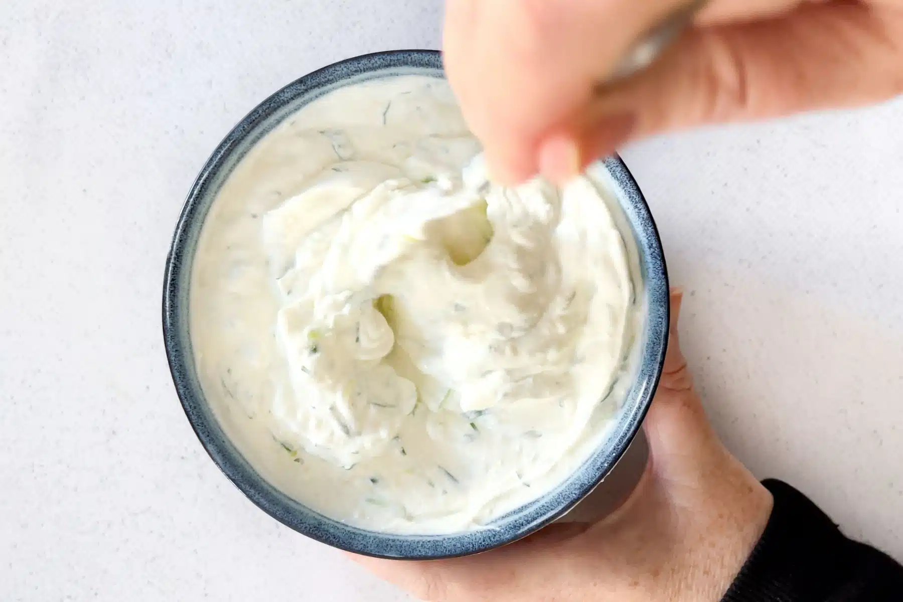 A white hand is stirring the tzatziki ingredients together in a blue bowl with a silver spoon.