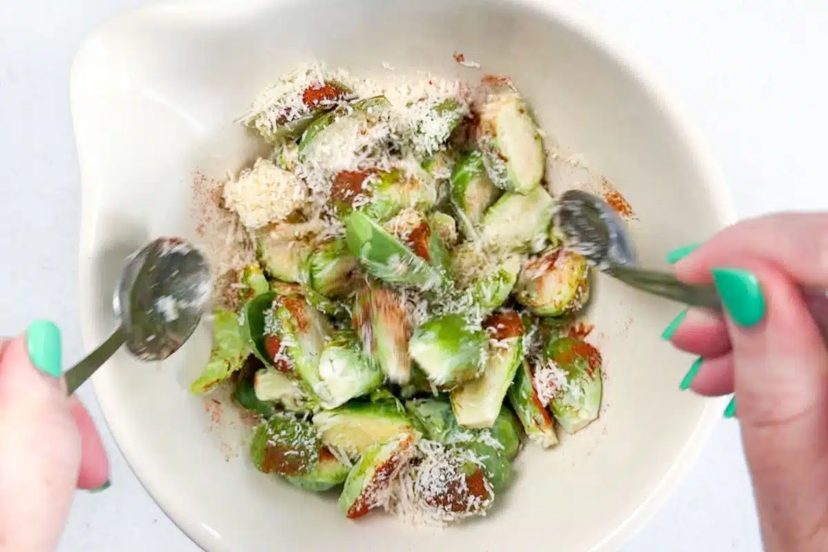Two white hands are tossing brussels sprouts, parmesan, oil, and spices in a large bowl.